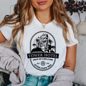 May include: White t-shirt featuring a black graphic of the Hollywood Tower Hotel. The graphic includes the text "The Hollywood Tower Hotel", "Drop In's Welcome", and "Established 1994".