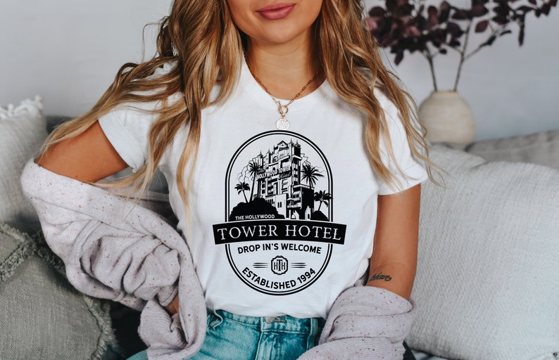May include: White t-shirt featuring a black graphic of the Hollywood Tower Hotel. The graphic includes the text "The Hollywood Tower Hotel", "Drop In's Welcome", and "Established 1994".