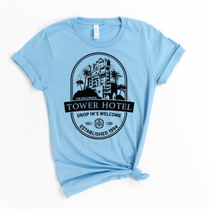 May include: Light blue t-shirt featuring a black graphic of the Hollywood Tower Hotel. The graphic includes the text "The Hollywood Tower Hotel", "Drop In's Welcome", and "Established 1994".