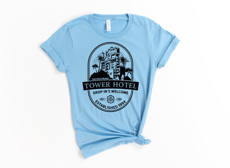 May include: Light blue t-shirt featuring a black graphic of the Hollywood Tower Hotel. The graphic includes the text "The Hollywood Tower Hotel", "Drop In's Welcome", and "Established 1994".