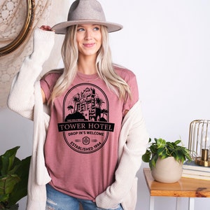 May include: A pink t-shirt with a black graphic of the Hollywood Tower Hotel, featuring the text "Tower Hotel", "Drop In's Welcome", and "Established 1994".