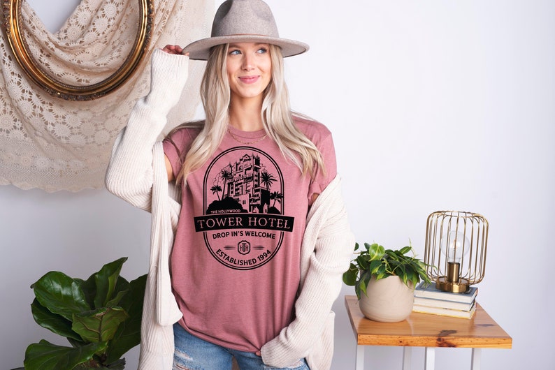 May include: A pink t-shirt with a black graphic of the Hollywood Tower Hotel, featuring the text "Tower Hotel", "Drop In's Welcome", and "Established 1994".