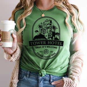 May include: A green t-shirt featuring a black graphic of the Hollywood Tower Hotel. The graphic includes the text "The Hollywood Tower Hotel", "Drop In's Welcome", and "Established 1994".