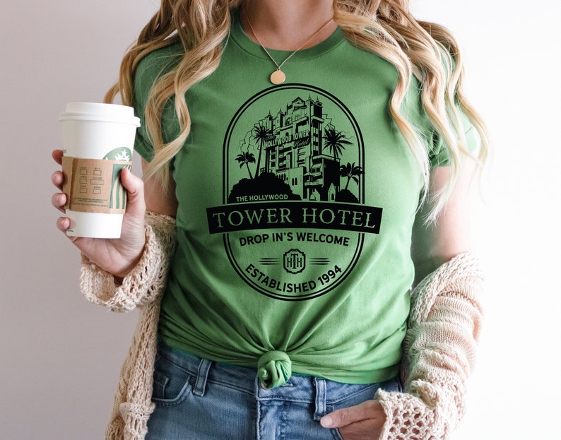 May include: A green t-shirt featuring a black graphic of the Hollywood Tower Hotel. The graphic includes the text "The Hollywood Tower Hotel", "Drop In's Welcome", and "Established 1994".