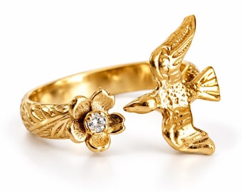 May include: A gold open-style ring featuring a textured band with a five-petaled flower on one end, centered with a small clear gemstone, and a bird in flight on the opposite end, both displaying intricate engraved detailing.