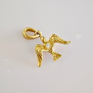 Op de afbeelding: Een gouden hanger in de vorm van een duif met een kleine, heldere edelsteen in het midden. De duif heeft gedetailleerde vleugels en een lus aan de bovenkant om aan een ketting te bevestigen.