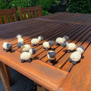 May include: A group of ten crocheted sheep figurines in white and gray are arranged on a wooden table. The sheep are all facing the same direction, with their heads tilted slightly upwards.