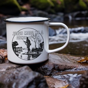 Peut inclure: Mug de camping en émail blanc avec un motif noir qui dit "Jésus a fait de moi un pêcheur d'hommes" avec une silhouette d'un homme pêchant et la référence biblique "Matthieu 4:19". Le graphique dit aussi "Je les attrape, il les nettoie".