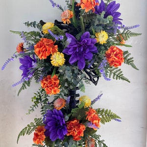 May include: A floral arrangement featuring purple, orange, and yellow flowers with green foliage. The flowers are arranged on a black metal base.