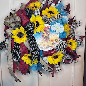 May include: A sunflower wreath with a red, black, and white checkered ribbon, a blue and white ribbon, and a sign that says "How Come Back".