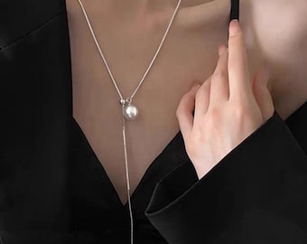 May include: A silver-toned necklace with a long, dangling design. The necklace features a large, round, pearl-like pendant and a delicate chain. The necklace is worn against a black top, creating a contrast that highlights the jewelry.