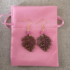 Pode incluir: Brincos de folha de madeira com ganchos dourados e contas rosa. São apresentados numa bolsa de cetim rosa, sobre um fundo neutro.