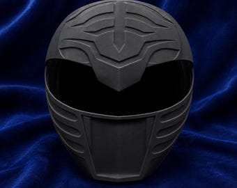 Op de afbeelding: Een matzwarte integraalhelm met een gestileerd, verhoogd geometrisch embleem op het voorhoofd en een brede, donkere vizieropening. De helm heeft een gladde, uniforme afwerking met gevormde horizontale ribbels langs de zijkanten en kin.