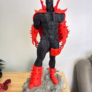 May include: A black and red figurine of a muscular character wearing a spiked cape, spiked gauntlets, and spiked boots. The figure stands on a grey, textured base featuring skull-like details, with a chain belt around the waist and a mask.