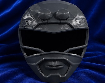 May include: A matte dark gray full-face helmet featuring a wide black visor, a sculpted chin section with defined lips, and two circular protrusions on the forehead area. The helmet has a textured surface with visible layered construction lines.