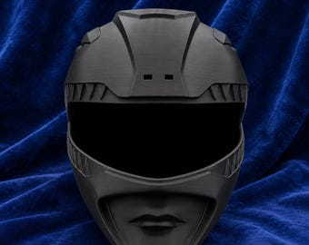 May include: A matte black full-face helmet featuring a stylized design with a wide visor opening, angular geometric panels on the crown, and a molded chin section that includes a subtle, raised lip-like detail at the very bottom center.