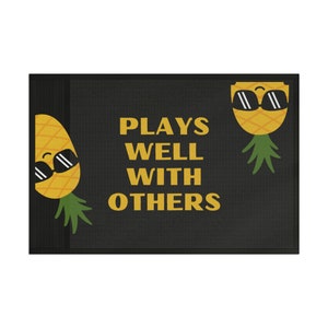 May include: A black doormat with the text "Plays Well With Others" in yellow letters. Two cartoon pineapples with sunglasses are on either side of the text.