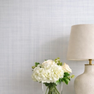 May include: A white nightstand with a drawer holds a cream-colored lamp with a beige shade and a bouquet of white flowers in a glass vase. The background is a light blue textured wall, and a white bed frame is partially visible.