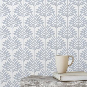 May include: A bedroom scene featuring a light gray and white patterned wallpaper with a repeating leaf design. A small, weathered wooden stool holds a mug and a book. A white bedspread and an open book on the floor complete the cozy setting.
