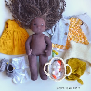 May include: A handmade cloth doll with brown skin and long, curly brown hair. The doll is surrounded by miniature clothing items, including a yellow dress, a blue jacket, a white hat, and gray shoes. A small, white basket holds a miniature felted baby.