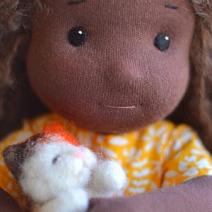 May include: A handmade doll with brown skin, black eyes, and brown hair. The doll is wearing a yellow dress with a white floral pattern and holding a small felted cat. The cat is white with brown and orange markings.