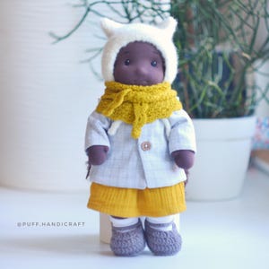 May include: A handmade doll wearing a white hooded hat, a yellow scarf, a light blue jacket, and yellow shorts. The doll also has gray crocheted shoes. The doll is standing on a white surface.