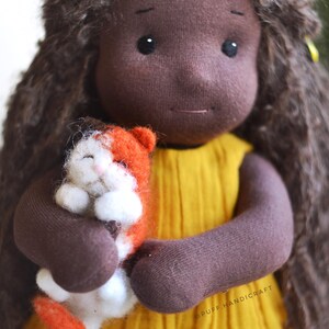 May include: A handmade doll with brown skin, long brown curly hair, and a yellow dress. The doll is holding a small orange and white cat. The doll's arms and hands are brown. The doll has black eyes and a small smile.