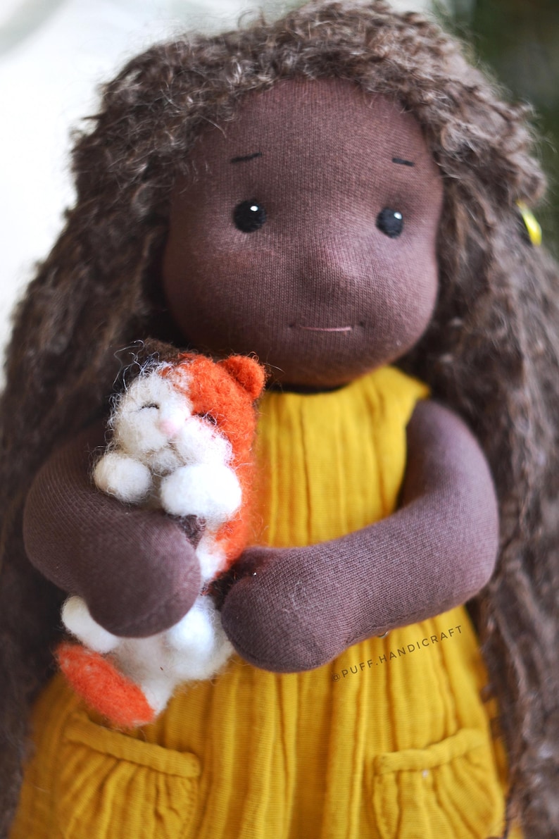 May include: A handmade doll with brown skin, long brown curly hair, and a yellow dress. The doll is holding a small orange and white cat. The doll's arms and hands are brown. The doll has black eyes and a small smile.
