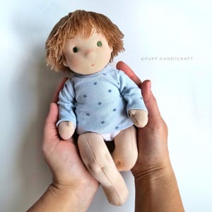 Puede incluir: Una mu&ntilde;eca de tela hecha a mano con cabello casta&ntilde;o, ojos verdes y una camisa azul con estrellas blancas. La mu&ntilde;eca lleva un pa&ntilde;al blanco y est&aacute; siendo sostenida en las manos de alguien.