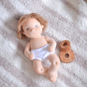 Puede incluir: Una mu&ntilde;eca de tela hecha a mano con cabello rubio y ojos azules que lleva un pa&ntilde;al blanco. La mu&ntilde;eca est&aacute; acostada sobre una manta de rayas blancas y marrones con un n&uacute;mero ocho de madera a su lado.