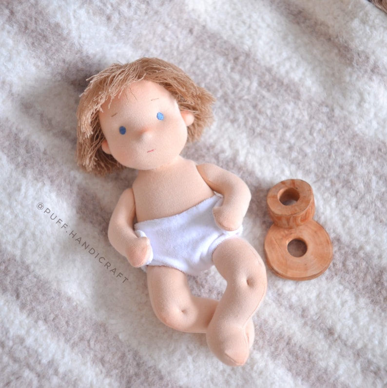 Puede incluir: Una mu&ntilde;eca de tela hecha a mano con cabello rubio y ojos azules que lleva un pa&ntilde;al blanco. La mu&ntilde;eca est&aacute; acostada sobre una manta de rayas blancas y marrones con un n&uacute;mero ocho de madera a su lado.