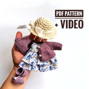Puede incluir: Un conjunto de muñeca en miniatura, que incluye un sombrero de paja con una cinta blanca, una rebeca lila, un vestido con estampado floral y zapatos a juego. El conjunto es para una muñeca y contiene el texto "PDF PATTERN + VIDEO".