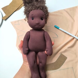 May include: A handmade cloth doll with dark brown skin and curly brown hair. The doll is wearing a dark purple outfit. The doll is being held in a person's hand. Sewing patterns and a marker are in the background.