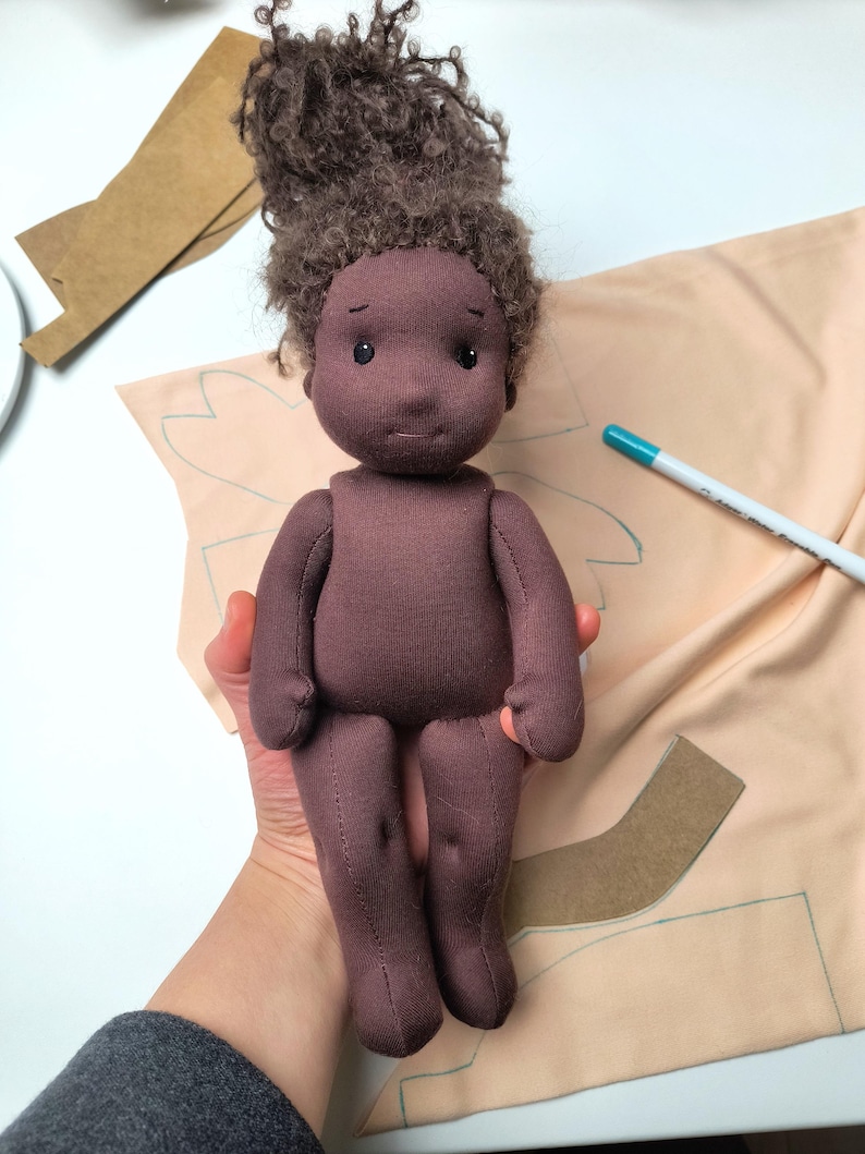 May include: A handmade cloth doll with dark brown skin and curly brown hair. The doll is wearing a dark purple outfit. The doll is being held in a person's hand. Sewing patterns and a marker are in the background.
