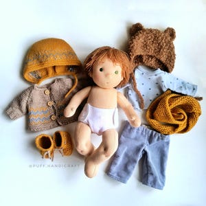 Puede incluir: Una mu&ntilde;eca hecha a mano con cabello casta&ntilde;o y ojos verdes que lleva una camisa a rayas azul y blanca, pantalones azules, un gorro de pelo marr&oacute;n, una bufanda de punto amarilla, un c&aacute;rdigan de punto amarillo y un gorro de punto amarillo. La mu&ntilde;eca tambi&eacute;n lleva un pa&ntilde;al blanco. La mu&ntilde;eca est&aacute; sentada sobre un fondo blanco. La imagen incluye el texto "@PUFF_HANDICRAFT".