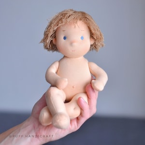 Puede incluir: Una mu&ntilde;eca de tela hecha a mano con cabello rubio y ojos azules. La mu&ntilde;eca est&aacute; sentada con las piernas cruzadas y tiene una expresi&oacute;n neutral.