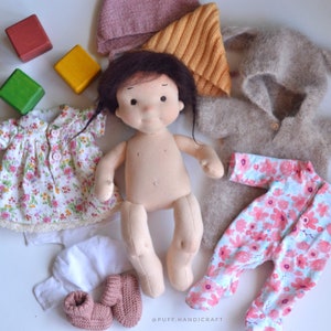 Puede incluir: Una mu&ntilde;eca desnuda con cabello casta&ntilde;o y un vestido floral, un gorro de punto rosa, un gorro de punto blanco, un par de botines de punto rosa, un mono floral rosa y blanco, una chaqueta de punto marr&oacute;n y un juego de bloques de madera de colores.