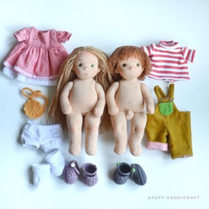 Puede incluir: Dos muñecas de tela con cabello de lana, una con cabello largo rubio y otra con cabello corto castaño. Los accesorios incluyen un vestido rosa con ojales, una camiseta a rayas rojas y blancas, un peto amarillo mostaza, pantalones cortos blancos, un bolso de ganchillo amarillo y dos pares de zapatos tejidos pequeños. El texto dice PUFF.HANDICRAFT.