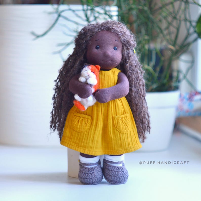 May include: A handmade doll with long brown curly hair, wearing a yellow dress and gray crocheted shoes. The doll is holding a small stuffed animal. The doll's skin is dark brown. The doll has a sweet, gentle expression.