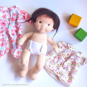 Puede incluir: Una mu&ntilde;eca de tela hecha a mano con cabello casta&ntilde;o que lleva un pa&ntilde;al blanco y un mono de estampado floral rosa. La mu&ntilde;eca est&aacute; rodeada de bloques de madera de colores y un vestido de estampado floral rosa.