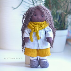 May include: A handmade cloth doll with long brown hair, wearing a yellow scarf, a light blue jacket, yellow shorts, and gray crocheted shoes. The doll has a neutral expression and is standing upright. The text "@PUFF.HANDICRAFT" is visible.