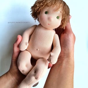 Puede incluir: Una mu&ntilde;eca de tela hecha a mano con pelo de lana marr&oacute;n, ojos verdes y una tez marr&oacute;n claro. La mu&ntilde;eca est&aacute; siendo sostenida por las manos de una persona.