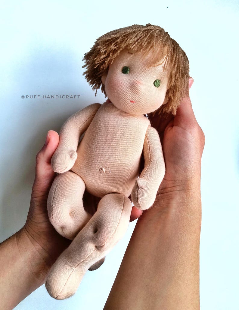Puede incluir: Una mu&ntilde;eca de tela hecha a mano con pelo de lana marr&oacute;n, ojos verdes y una tez marr&oacute;n claro. La mu&ntilde;eca est&aacute; siendo sostenida por las manos de una persona.