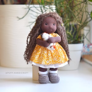 May include: A handmade doll with dark skin, long brown hair, and a yellow floral dress. The doll is holding a small white stuffed animal. It is wearing gray crocheted shoes and white socks. The doll is approximately 10 inches tall.