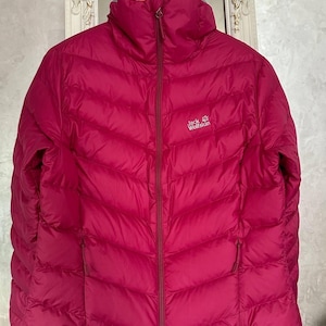 Könnte beinhalten: Eine magentafarbene, gesteppte Pufferjacke mit durchgehendem Reißverschluss und hohem Kragen. Das Logo "Jack Wolfskin" ist auf der Brust zu sehen. Die Jacke hat zwei Reißverschlusstaschen und hängt an einem Holzbügel.