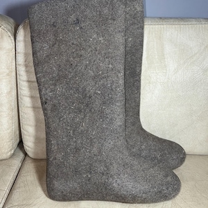 Puede incluir: Un par de botas de fieltro grises. Las botas son altas, llegando hasta la mitad de la pantorrilla. El material tiene un aspecto texturizado. Las botas están sobre una superficie de color claro.