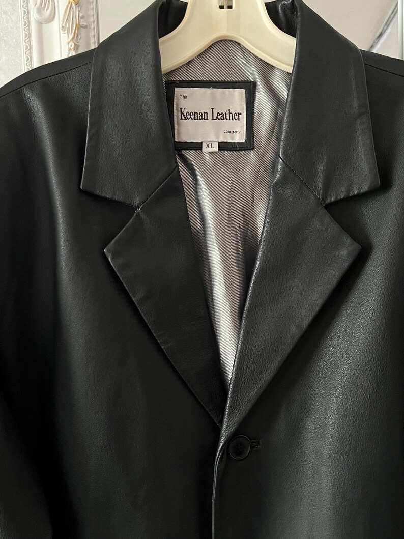 Peut inclure: Un blazer en cuir noir avec un revers crant&eacute; et une fermeture &agrave; un seul bouton. La doublure int&eacute;rieure est de couleur gris clair. L'&eacute;tiquette indique "The Keenan Leather company" et la taille est XL.