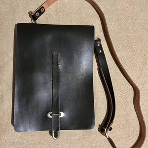 May include: A rectangular black leather bag featuring a vertical strap closure with a silver-toned metal buckle. The bag includes a long, adjustable shoulder strap made of two-toned brown and black leather, attached with silver metal hardware.