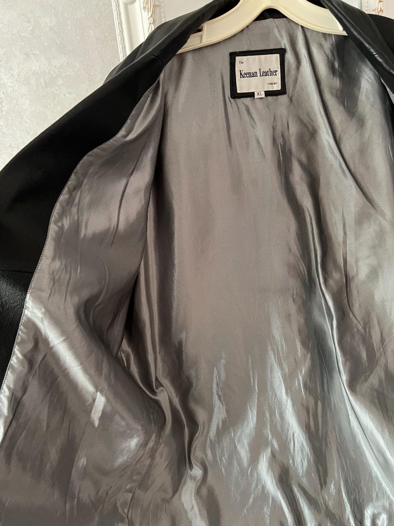 Peut inclure: Veste en cuir noir avec une doublure en satin gris. La veste est ouverte, r&eacute;v&eacute;lant la doublure int&eacute;rieure. Une &eacute;tiquette &agrave; l'int&eacute;rieur de la veste indique "Keenan Leather". La veste est suspendue &agrave; un cintre blanc.