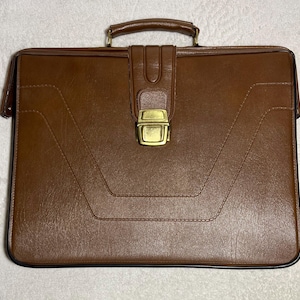May include: A brown leather briefcase with a gold-colored clasp and handle. The briefcase has a rectangular shape with stitched detailing and a secure closure. The overall design is classic and professional, suitable for carrying documents and essentials.